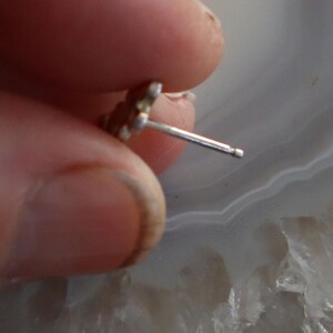 May include: A small, silver-colored stud earring with a floral design. The earring post is visible, and the earring is held by a person's fingers. The background is a light gray and white marbled surface.