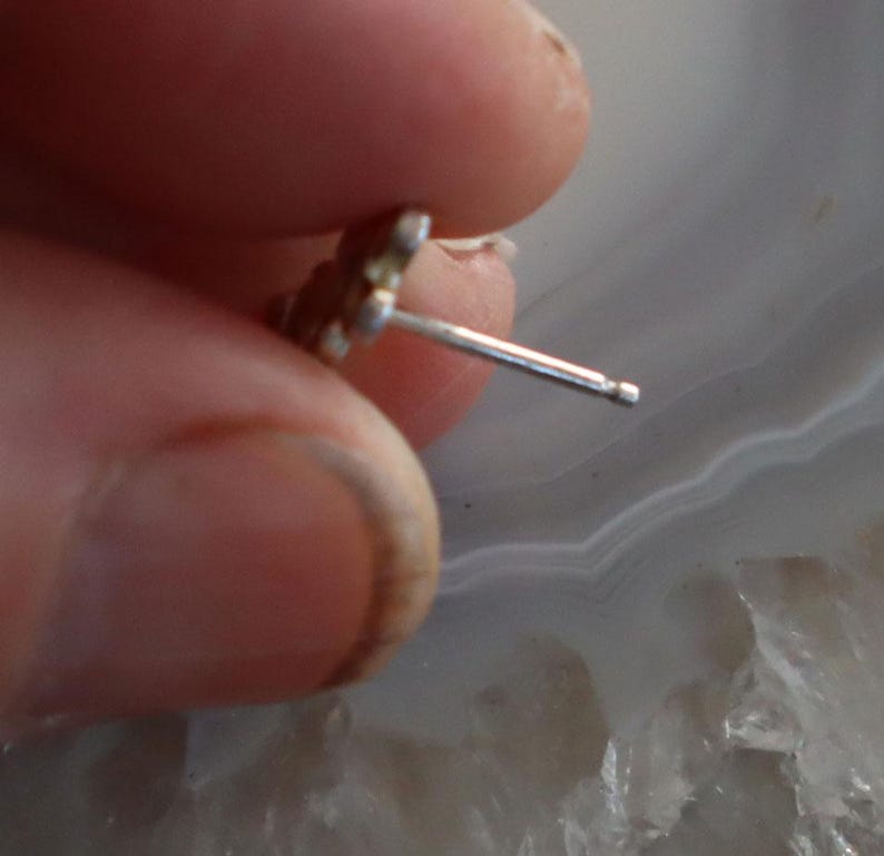 May include: A small, silver-colored stud earring with a floral design. The earring post is visible, and the earring is held by a person's fingers. The background is a light gray and white marbled surface.