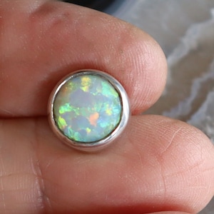 Op de afbeelding: Een ronde, iriserende opaal edelsteen gezet in een zilveren bezel. De opaal toont een kleurenspel met flitsen van groen, oranje en blauw. De edelsteen wordt in een hand gehouden.