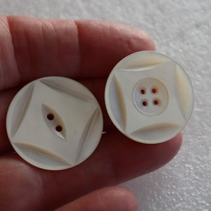Può includere: Due bottoni rotondi bianco sporco con un design quadrato. Un bottone ha due fori e l'altro quattro fori. I bottoni sono tenuti in una mano.