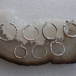 May include: A collection of seven silver-toned hoop earrings of varying sizes and designs. Some hoops feature twisted or textured metal patterns, whilst others are smooth. Each earring includes a secure clasp mechanism for attachment to the earlobe.