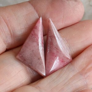 May include: Two triangular, pink gemstones with a faceted design. The gemstones have a marbled appearance with darker pink and white veining. The stones are held in a hand, showcasing their shape and color.