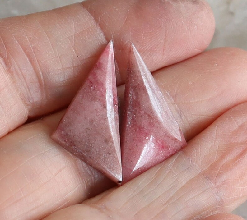 May include: Two triangular, pink gemstones with a faceted design. The gemstones have a marbled appearance with darker pink and white veining. The stones are held in a hand, showcasing their shape and color.
