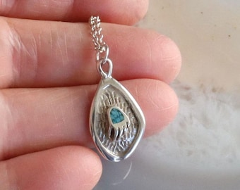 May include: Silver pendant necklace with a teardrop-shaped pendant. The pendant features a textured design with a turquoise-colored inlay in the center. The necklace has a delicate chain.