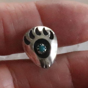 May include: A silver-toned bear paw-shaped stud earring with a turquoise-colored stone in the center. The earring has a black border around the stone and the paw pads.