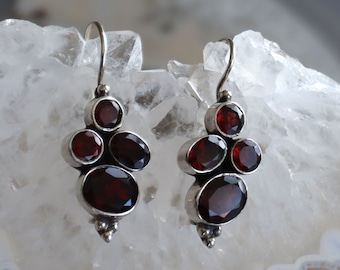 May include: A pair of silver earrings featuring multiple oval and round, deep red gemstones. The earrings have a cluster design with a hook closure. The gemstones are set in a silver metal setting, creating a striking contrast.