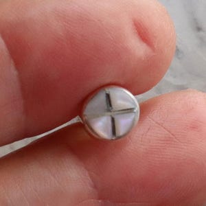 May include: A small, round earring with a silver setting. The earring features a cross design inlaid with iridescent mother-of-pearl. The earring is held between two fingers.