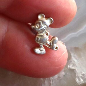 May include: A small, silver-colored charm in the shape of Mickey Mouse. The charm is detailed with visible ears, a smiling face, and iconic shorts. The figure is in a walking pose, with one leg extended.