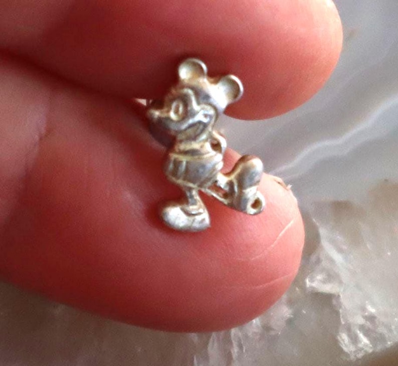 May include: A small, silver-colored charm in the shape of Mickey Mouse. The charm is detailed with visible ears, a smiling face, and iconic shorts. The figure is in a walking pose, with one leg extended.