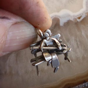 May include: A silver-toned pendant featuring an angel seated on a bench. The angel has wings and is holding a musical instrument. The bench has a slatted design. The pendant is held by a finger.