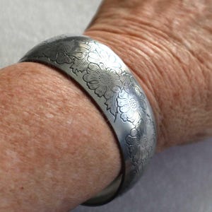 May include: A wide silver-toned metal cuff bracelet featuring an engraved floral pattern with multiple detailed flowers and leaves covering the outer surface. The bracelet has a smooth, curved profile and a polished finish that reflects light.