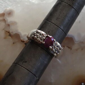 May include: A silver ring with a large, oval, deep red gemstone at the center. The ring has a textured design on either side of the gemstone, with small, round silver accents. The ring is displayed on a dark metal ring sizer.