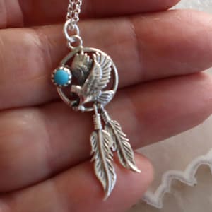 May include: A silver-toned necklace pendant featuring a circular dreamcatcher frame with an eagle in the center, a small round turquoise-colored stone on the left side, and two dangling silver-toned feathers hanging from the bottom of the frame.