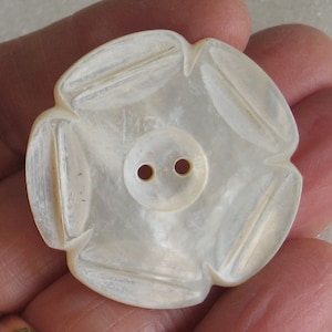 May include: A pearlescent, flower-shaped button with a central, smaller button featuring two holes. The button has a carved design on each petal, creating a textured appearance. The button is likely made of mother-of-pearl.