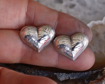 May include: Two silver heart-shaped pendants with detailed engraved designs. The pendants have a textured border and intricate patterns on the surface. The hearts are displayed against a neutral background.