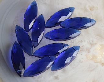 May include: A collection of faceted, marquise-shaped, sapphire-blue glass beads. The beads are arranged on a white and gray marbled surface, showcasing their geometric cuts and vibrant color. These beads are suitable for jewelry making.