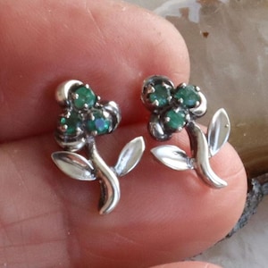 Könnte beinhalten: Ein Paar silberfarbene Ohrstecker in Blumenform. Jeder Ohrring hat ein Blumendesign mit drei grünen Edelsteinen in der Mitte und zwei Blättern. Die Ohrringe sind klein und zart, geeignet für den täglichen Gebrauch.