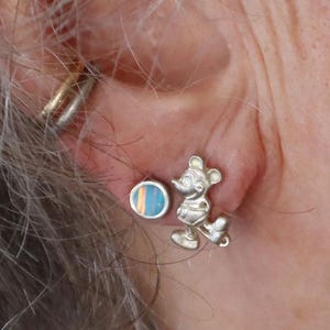 May include: A pair of silver earrings. One earring is a small, round stud with blue, orange, and yellow stripes. The other earring is a silver Mickey Mouse figure in a standing pose. The earrings are worn on an ear.