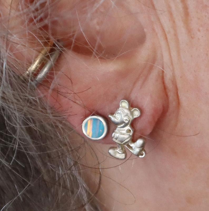 May include: A pair of silver earrings. One earring is a small, round stud with blue, orange, and yellow stripes. The other earring is a silver Mickey Mouse figure in a standing pose. The earrings are worn on an ear.