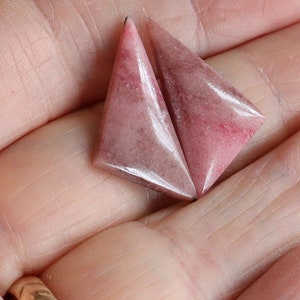 May include: Two triangular, pink gemstones with a polished surface. The stones have a gradient of color, with darker pink hues at the edges and lighter shades in the center. The gemstones are held in a hand.