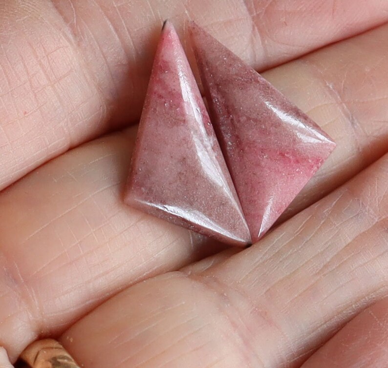 May include: Two triangular, pink gemstones with a polished surface. The stones have a gradient of color, with darker pink hues at the edges and lighter shades in the center. The gemstones are held in a hand.