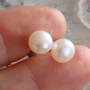 Op de afbeelding: Twee ronde, witte parels met een glanzende glans. De parels worden tussen vingers gehouden, waardoor hun gladde, bolvormige vorm en subtiele iriserende effect worden benadrukt. Deze parels zijn een klassiek sieraad.