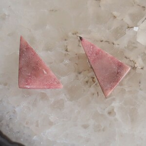 May include: Two triangular, pink gemstones with a mottled appearance. The gemstones are set against a white and beige background, showcasing their natural color variations and textures. These stones are likely for jewelry making.