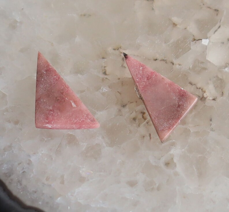 May include: Two triangular, pink gemstones with a mottled appearance. The gemstones are set against a white and beige background, showcasing their natural color variations and textures. These stones are likely for jewelry making.