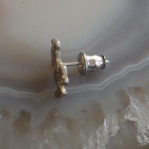 May include: A silver-colored stud earring with a decorative design. The earring post and backing are visible. The earring is set against a light-colored, patterned background.