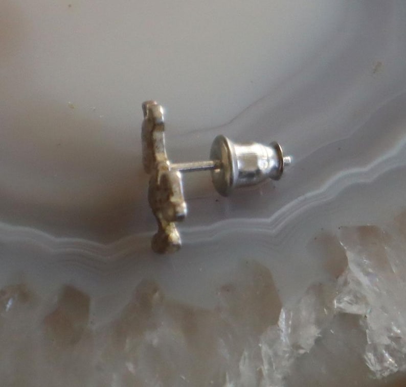 May include: A silver-colored stud earring with a decorative design. The earring post and backing are visible. The earring is set against a light-colored, patterned background.