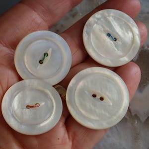 May include: Four round, iridescent white buttons with colorful thread. The buttons have a pearlescent sheen and a classic two-hole or four-hole design. The thread colors are green, blue, and orange.