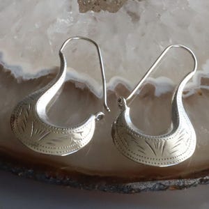 May include: A pair of silver-toned earrings with a curved, crescent-like design. The earrings feature etched patterns along the curved surface, with a hook for securing them to the ear. The earrings are displayed on a light-colored, patterned surface.