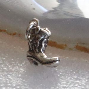 May include: A small, silver-colored charm in the shape of a cowboy boot. The boot has intricate detailing, including a pointed toe, a curved heel, and decorative stitching. The charm is designed to be attached to a bracelet or necklace.