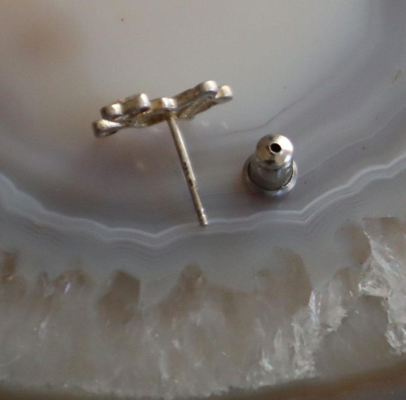 May include: A silver-colored stud earring with a floral design, next to a matching earring back. The earring has a post for insertion into the ear. The background is a light gray and white marbled surface.