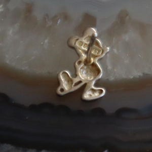 May include: A gold-colored Mickey Mouse charm. The charm is in the shape of Mickey Mouse, with his iconic silhouette. The charm has a loop at the top for attaching to a bracelet or necklace. The word "Disney" is visible on the charm.