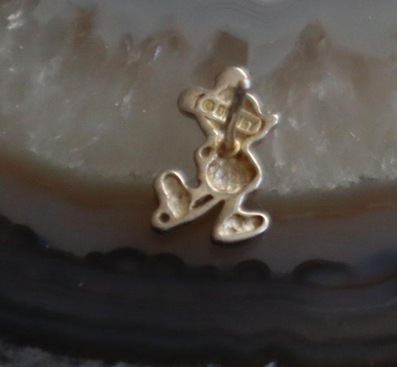 May include: A gold-colored Mickey Mouse charm. The charm is in the shape of Mickey Mouse, with his iconic silhouette. The charm has a loop at the top for attaching to a bracelet or necklace. The word "Disney" is visible on the charm.