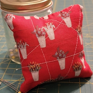 May include: A red pincushion with a floral pattern and white stitching. The pincushion is square-shaped and features a repeating design of white flower pots. A glass jar with a gold lid and sewing pins is in the background.