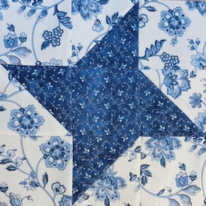 Op de afbeelding: Een vierkant quiltblok met een sterpatroon. De ster is gemaakt van een donkerblauwe bloemenstof, afgezet tegen een witte stoffen achtergrond met een blauw bloemenpatroon. Het blok is een patchwork-ontwerp.