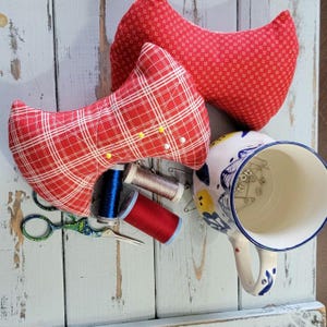 May include: A collection of sewing supplies on a distressed, light blue wooden surface. Includes two red pin cushions, spools of thread, decorative scissors, and a floral-decorated mug filled with pins. The pin cushions have plaid and floral patterns.
