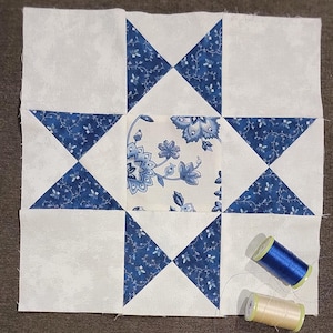May include: A square quilt block with a star design. The block features white and blue floral patterned fabric. The star points are made of blue fabric with a white floral design. Two spools of thread, one blue and one beige, are next to the block.
