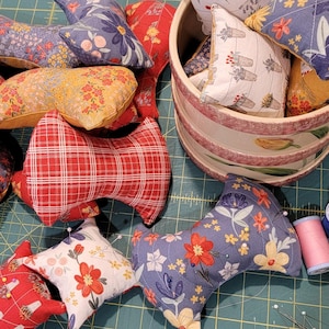 May include: A collection of colorful fabric pincushions in various patterns, including floral, plaid, and geometric designs. The pincushions are arranged on a green cutting mat with spools of thread and sewing pins.