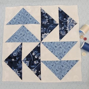 May include: A square quilt block with a geometric design. The block features alternating white and blue triangles, some with floral patterns. Spools of thread and buttons are arranged nearby, suggesting a sewing project.