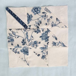 Può includere: Un blocco patchwork con sfondo color crema e motivi floreali blu. Il blocco è composto da diversi pezzi di tessuto, tra cui una striscia diagonale e triangoli. Il design presenta un motivo floreale classico.