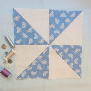 Peut inclure: Un bloc de patchwork carré avec un motif de moulinet. Il est composé de tissu bleu clair orné de plumes blanches et de tissu blanc cassé à pois. Des fils à coudre et des boutons sont disposés autour du bloc, évoquant un projet de couture.