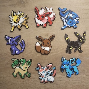 Puede incluir: Nueve figuras de pixel art coloridas de varios personajes de Pokémon, incluyendo Jolteon, Flareon, Vaporeon, Espeon, Eevee, Umbreon, Leafeon, Sylveon y Glaceon. Cada figura está hecha de pequeñas cuentas de colores.