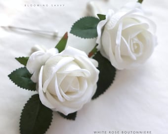 May include: Two white rose boutonnieres with green leaves and stems. The roses are made of fabric and have a realistic appearance. The text "BLOOMING SAVVY" and "WHITE ROSE  BOUTONNIERE" is visible in the image.