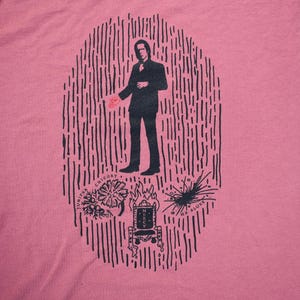 Puede incluir: Una camiseta rosa con un estampado gráfico en blanco y negro de un hombre de pie dentro de un círculo de líneas. El gráfico también incluye un trono con la palabra "misericordia" en él, una flor con la palabra "achicoria" al lado y una estrella con las palabras "ha blues" al lado.