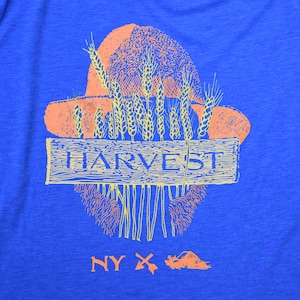 Puede incluir: Una camiseta azul con un gráfico de un sombrero de paja con tallos de trigo y un letrero de madera que dice "HARVEST". El gráfico es de color naranja y amarillo. La parte inferior de la camiseta tiene el texto "NY X" en naranja.