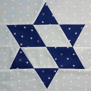 May include: A close-up of a fabric block featuring a large blue star with white star accents. The star is set against a light gray background with white polka dots. The design is a geometric pattern.