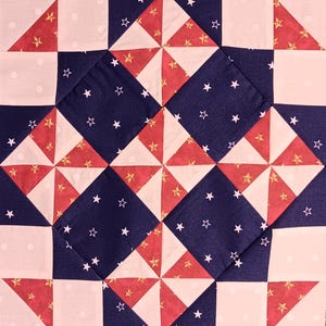 Op de afbeelding: Close-up van een quiltblok met een patriottisch thema. Het ontwerp toont afwisselende vierkanten van wit met kleine stippen, rode driehoeken met gouden sterren en marineblauwe vierkanten met witte sterren. De quilt is gemaakt van stof.